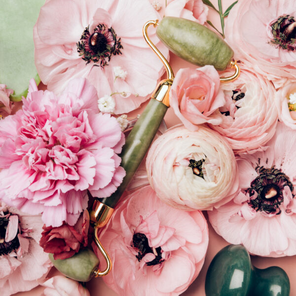 Jade face rollers and Gua sha stones in ranunculus flowers in flatlay on pink background with copy space. Bunch of Persian buttercup in floral arrangments, top view